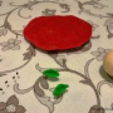 Modele uma bola com a massa branca, um círculo achatado com a vermelha e faça folhinhas com a verde. Precisaremos, ainda, de grãos de gergelim preto e de cravos.
